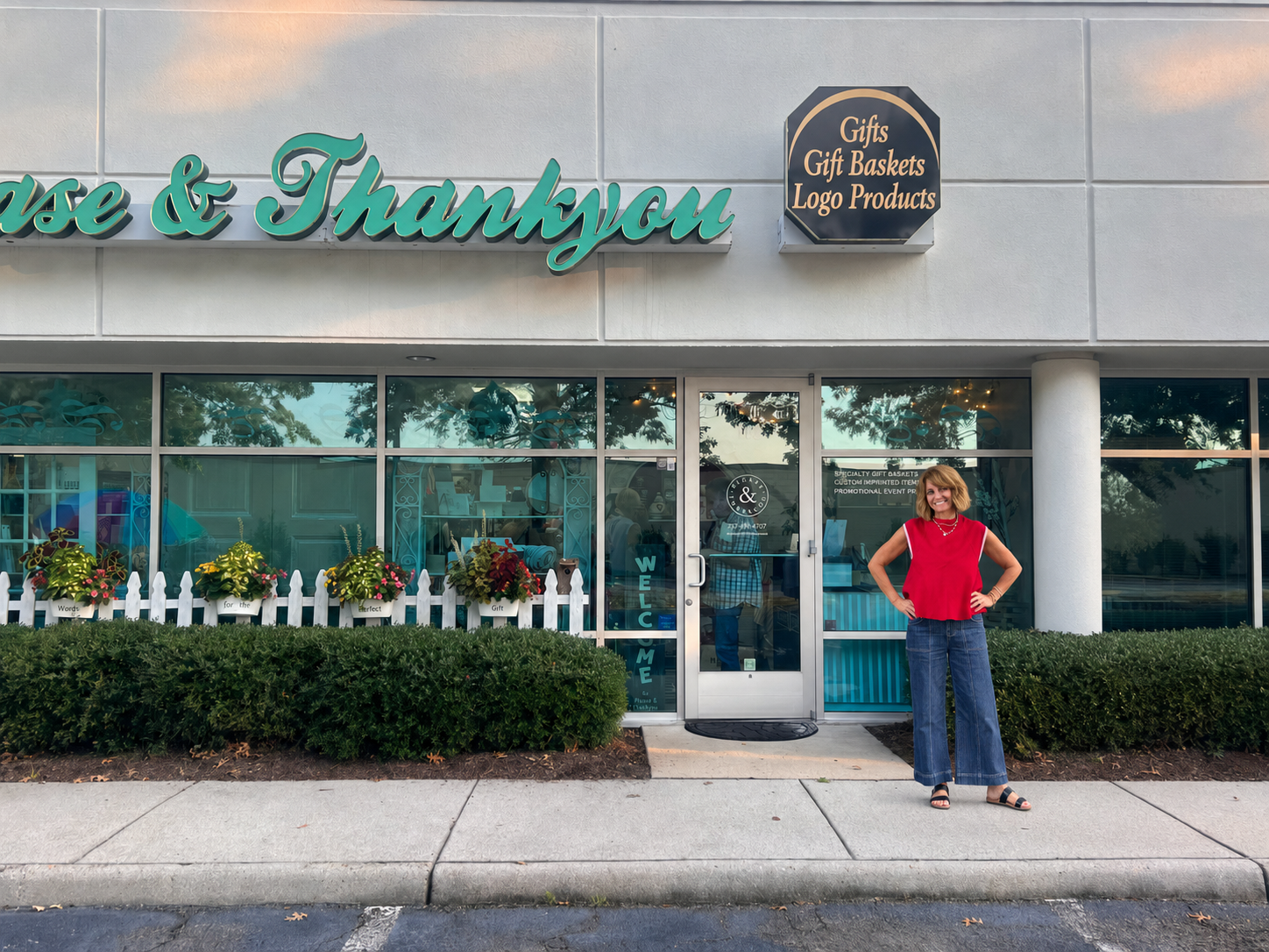 A woman stands on the sidewalk in front of a storefront building with large windows, a gray facade, and teal lettering that reads 'Case & Thankyou.' A black sign above the entrance reads 'Gifts Gift Baskets Logo Products.